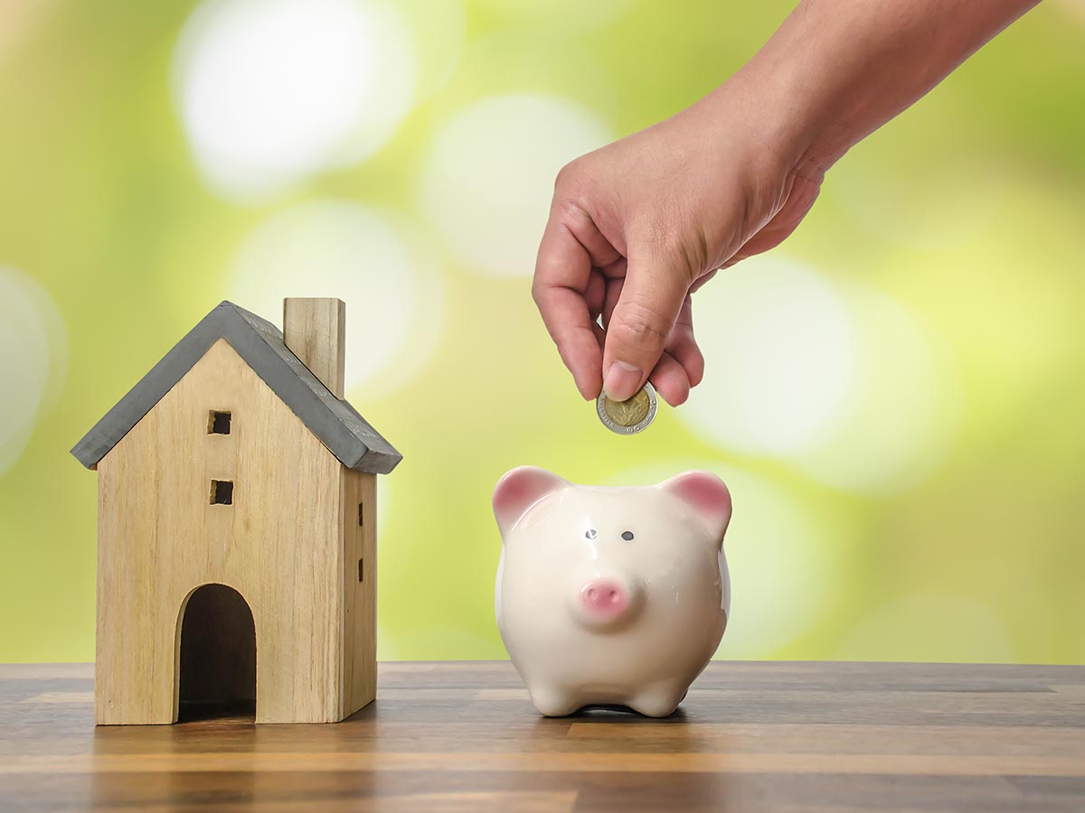 Person putting money into a piggy bank next to a wooden model of a house